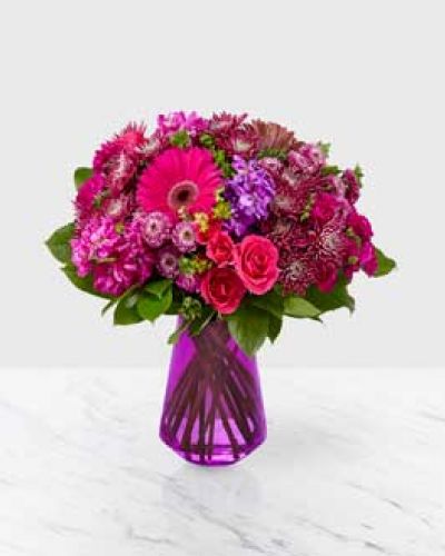 <p>Our Blushing Bouquet is sure to turn heads with its explosion of vibrant purple and hot pink tones. There’s no comparison to the stunning array of gerberas, mini carnations, spray roses, button poms, and stock, complemented by touches of salal, placed in a vibrant purple glass vase.</p>